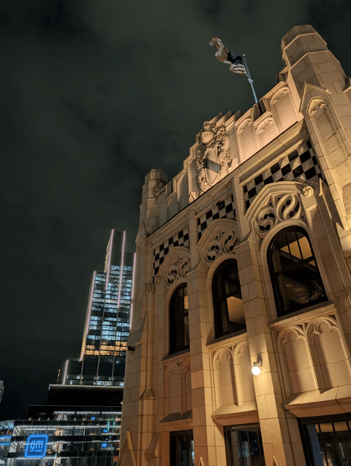 A Gothic building lit at night with a United States flag and the GM Renaissance Center glowing behind