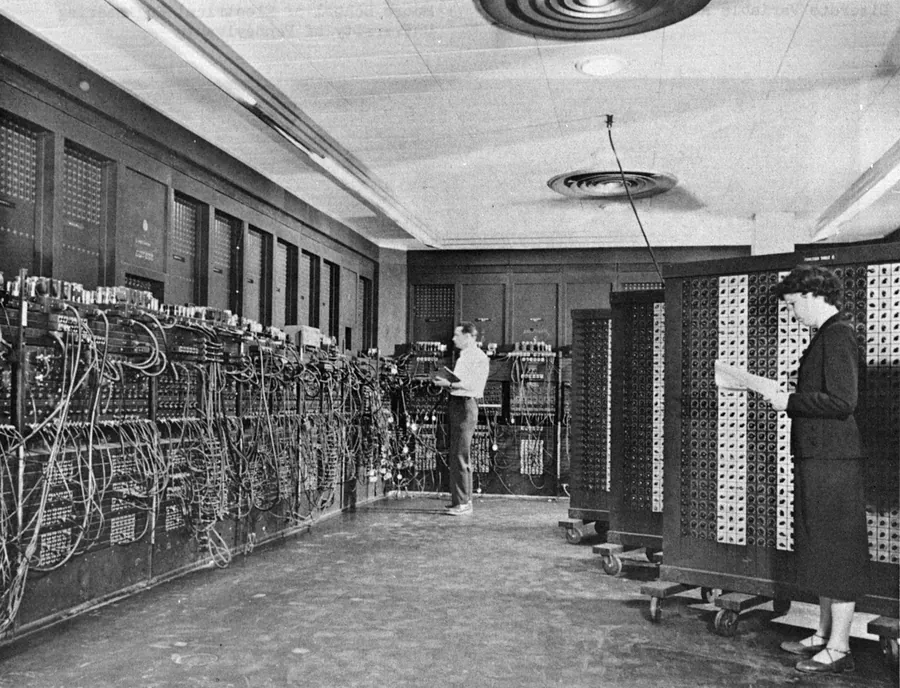 A long room lined on both sides with the ENIAC's tall cabinets and cable panels, with two operators standing at the units holding sheets of paper.